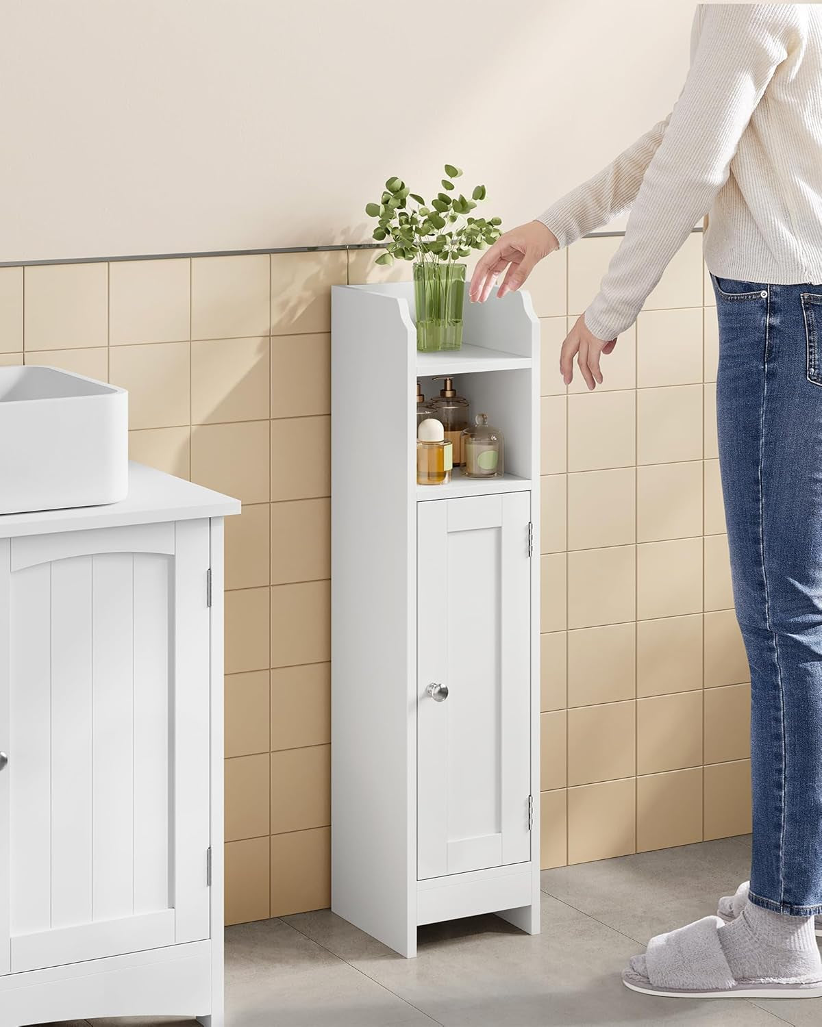 Narrow white floor cabinet beside a sink with toiletries on open shelves and a person reaching toward the top