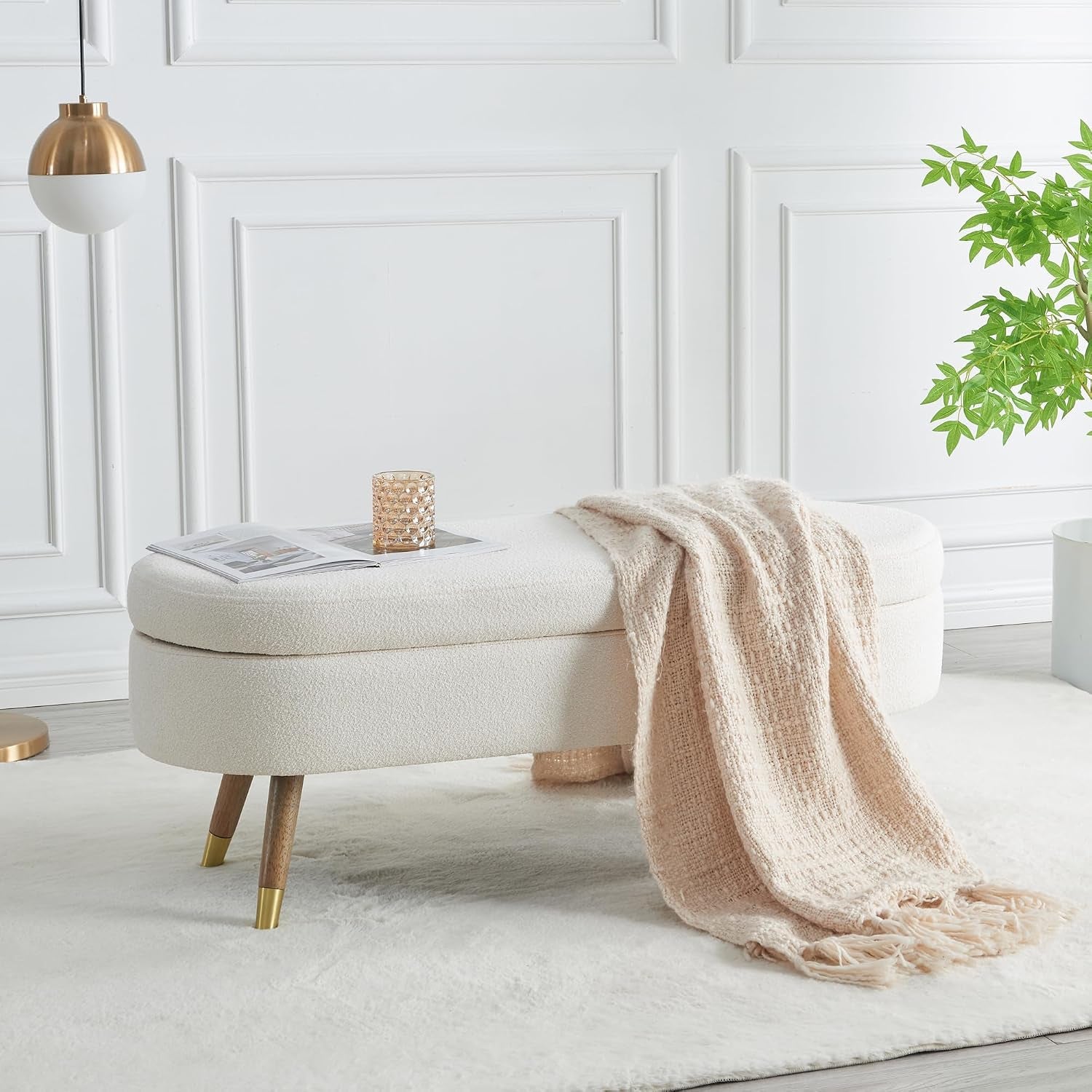 Oval cream boucle upholstered storage bench with gold-tipped wooden legs in a bright room.