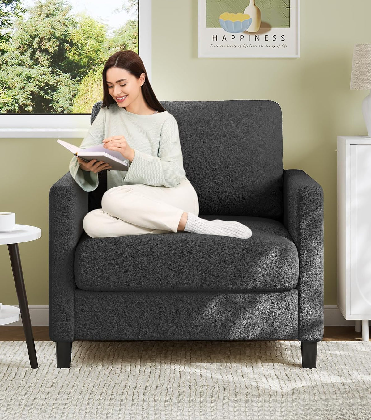 Front close view of a charcoal-gray small loveseat with a woman reading, showing straight arms and plush cushions.