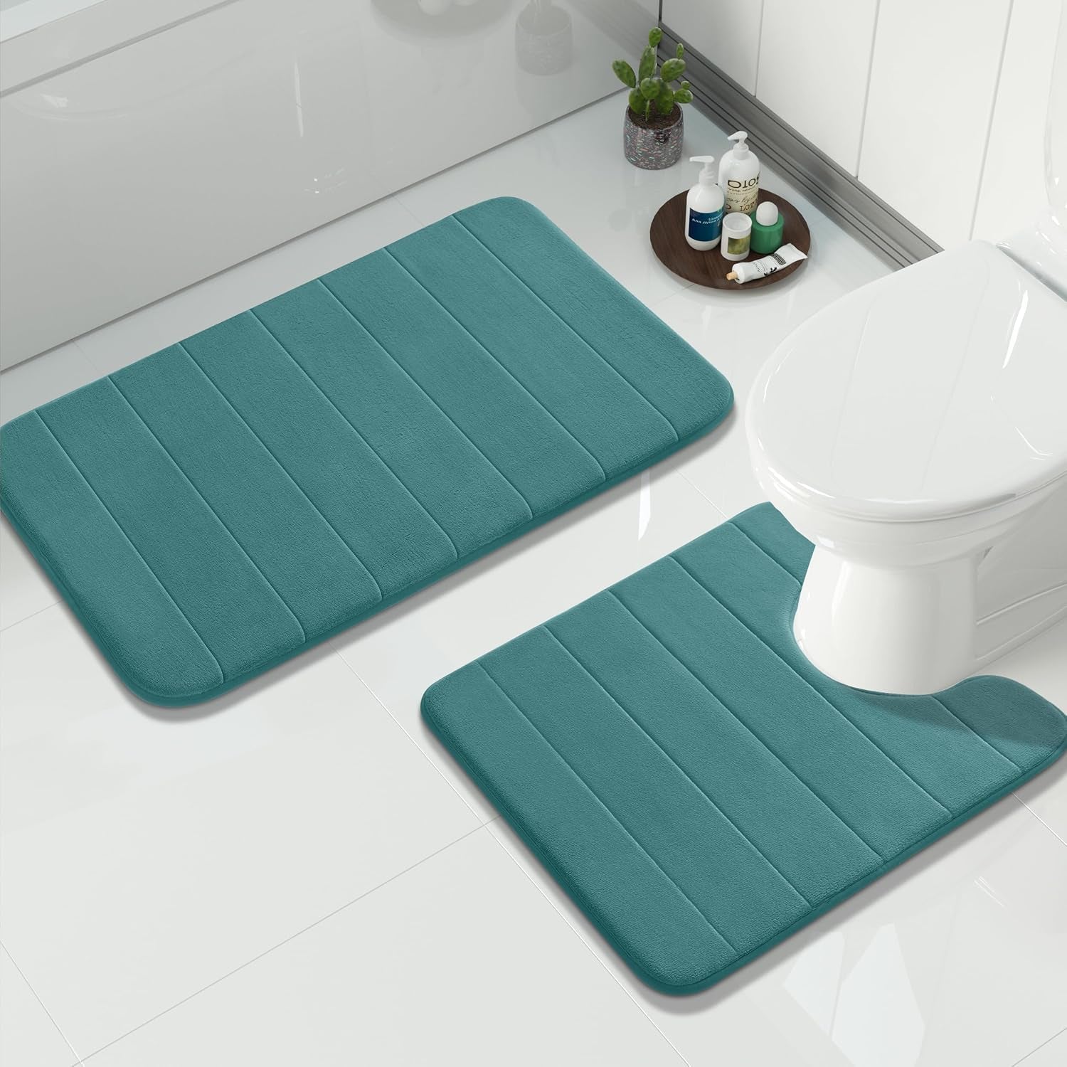 Large rectangular bath mat and a U-shaped toilet rug in blue-green on shiny white tile with toiletries on a tray.