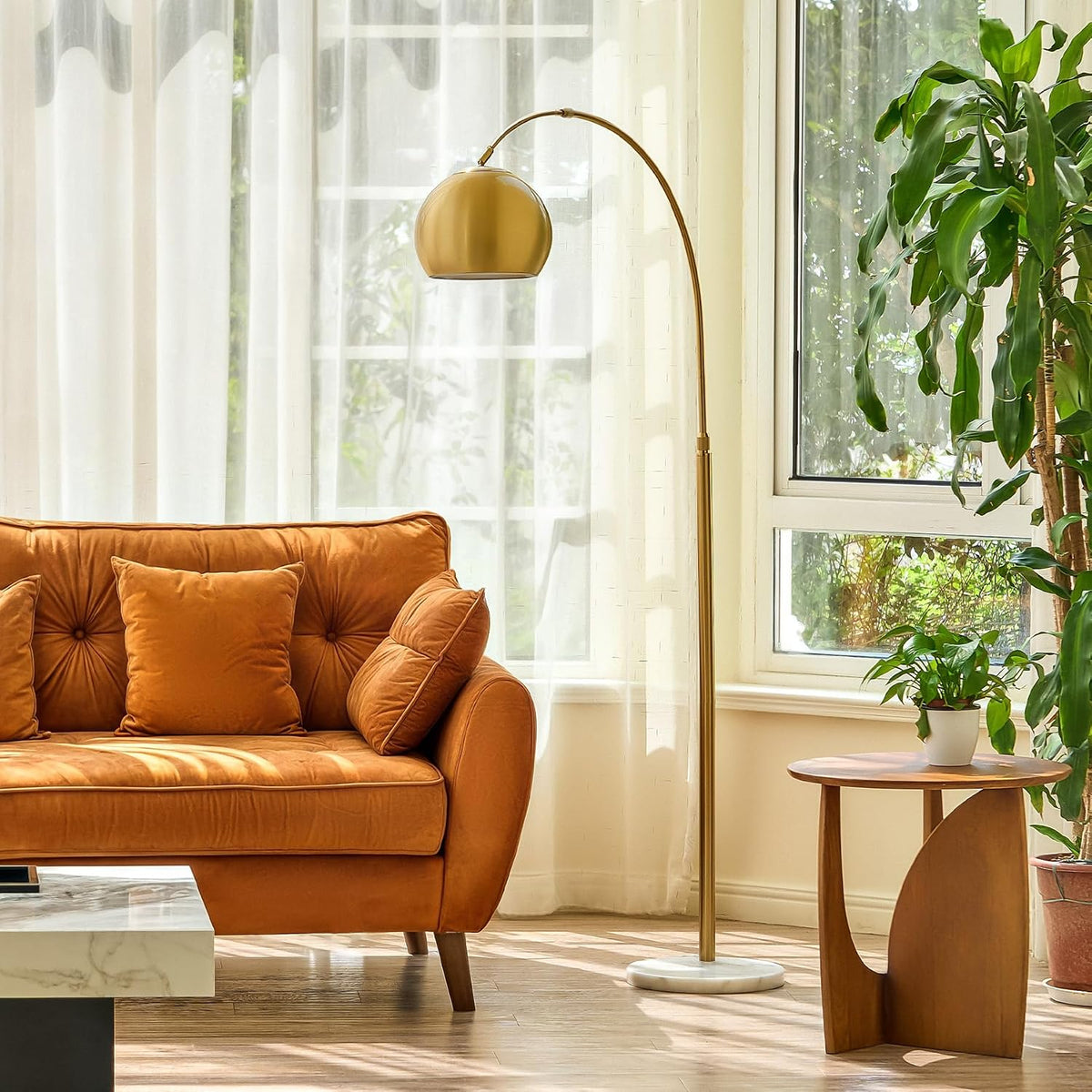 Gold arc floor lamp standing beside an orange tufted sofa and wooden side table by a sunny window