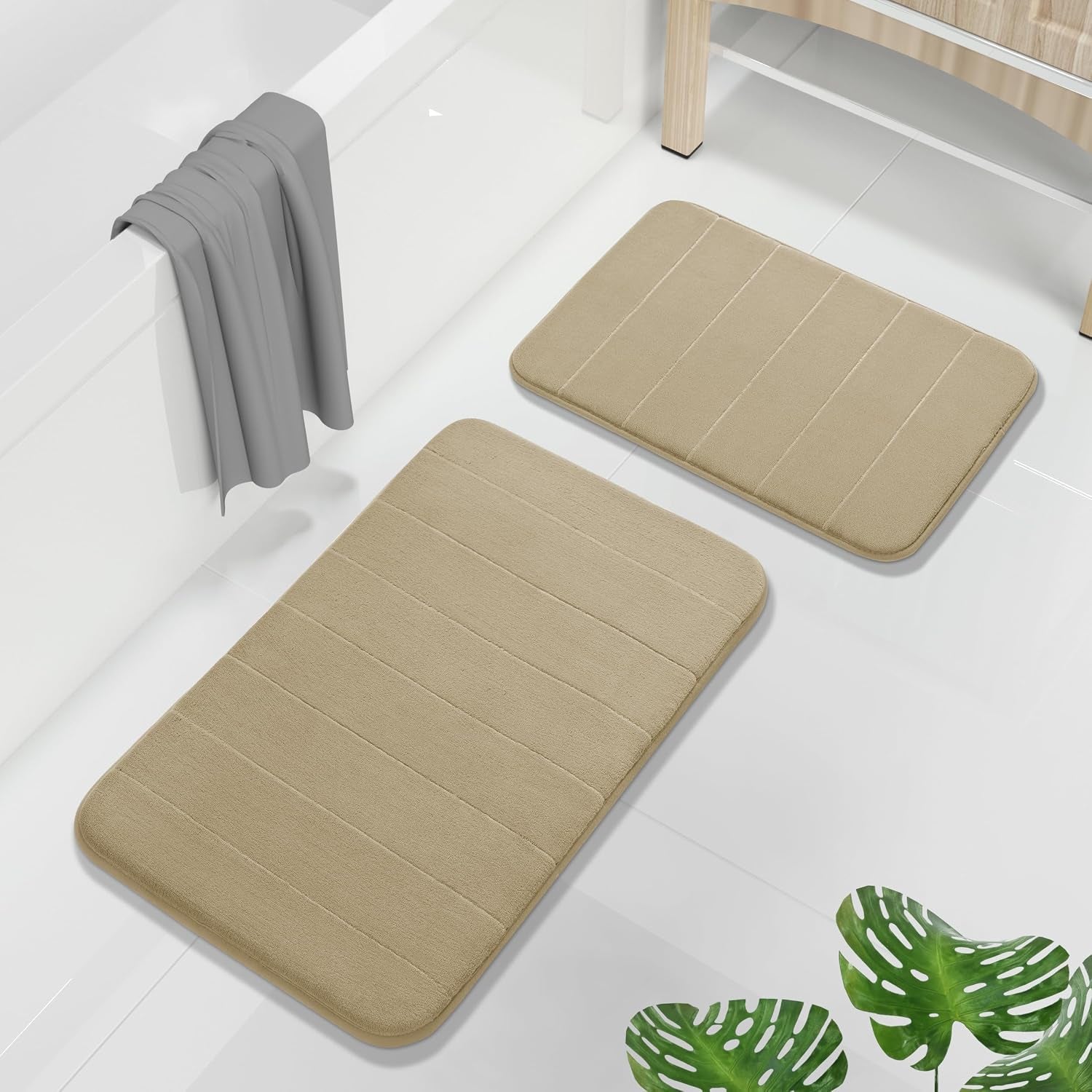 Beige two-piece bath mat set on white tiled floor beside a bathtub and wooden storage bench