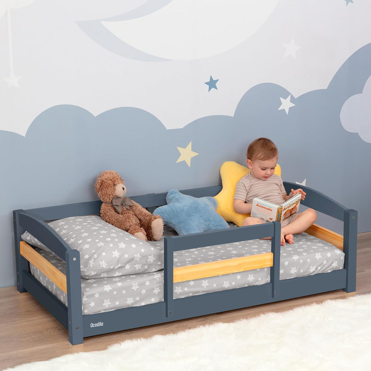 Low gray toddler bed with wooden accent rails, stuffed toys and a child reading in a star-decorated room.