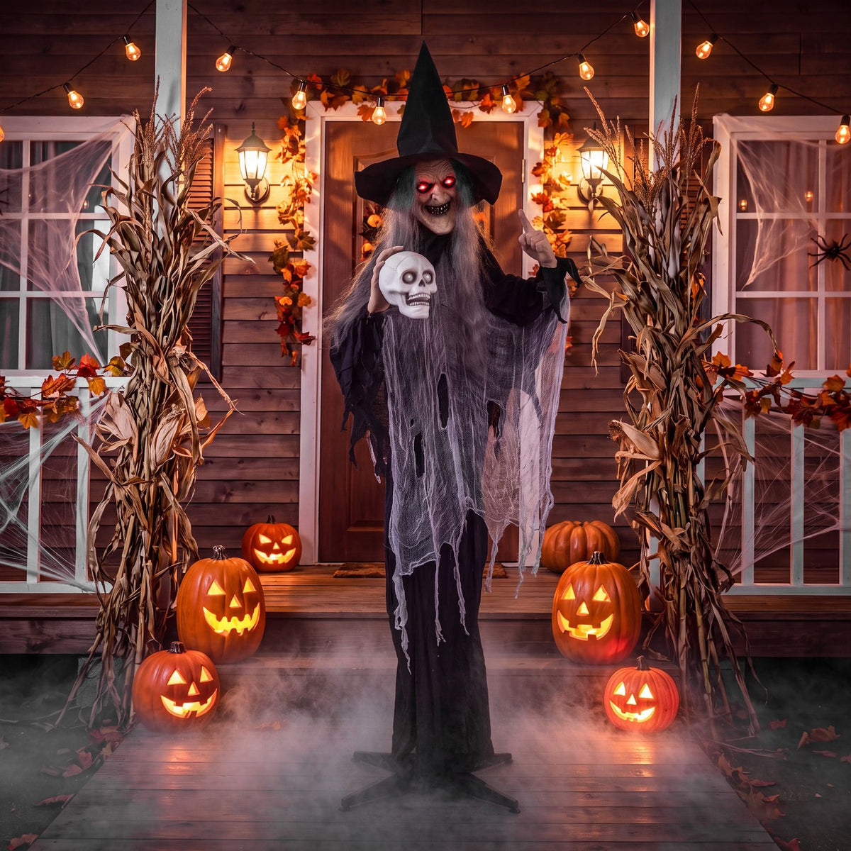A skull-holding witch stands on a decorated porch among glowing jack-o'-lanterns and dried cornstalks.