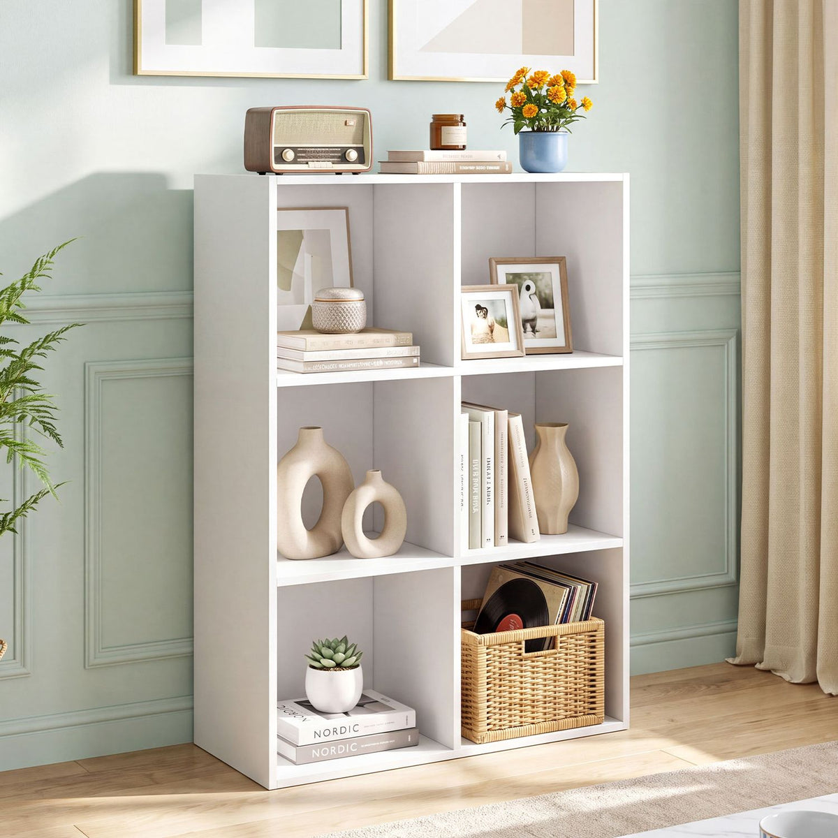 White six-cubby bookcase styled with books, framed photos, vases, and a woven basket in a pale green room.