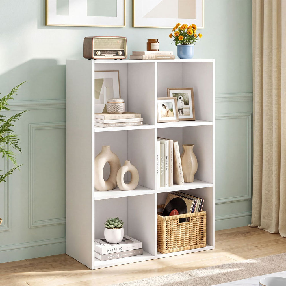 White six-cubby bookcase styled with books, framed photos, vases, and a woven basket in a pale green room.
