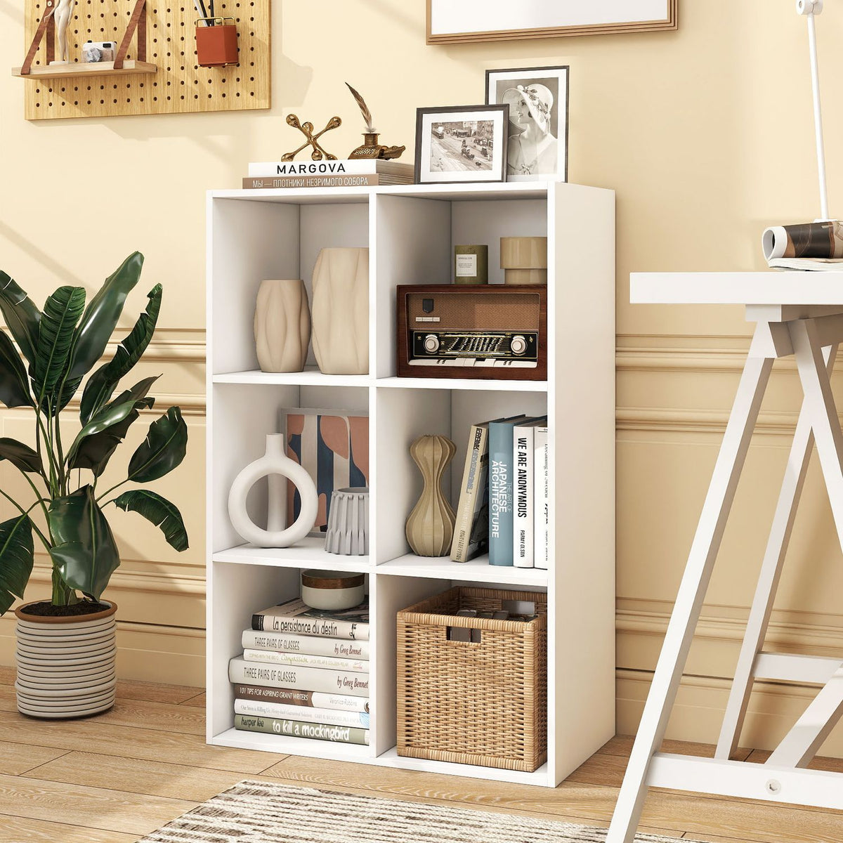 White cube bookcase filled with decor and books, beside a leafy plant and desk in a warm-toned room.