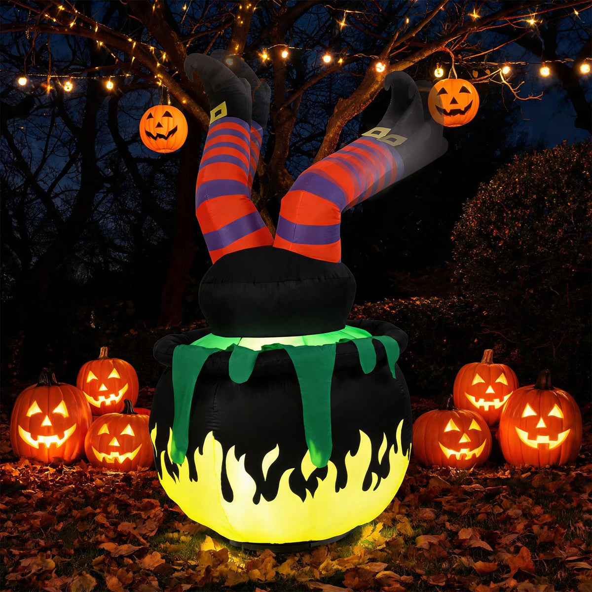 Glowing cauldron with striped witch legs displayed among pumpkins beneath string lights in a wooded yard.