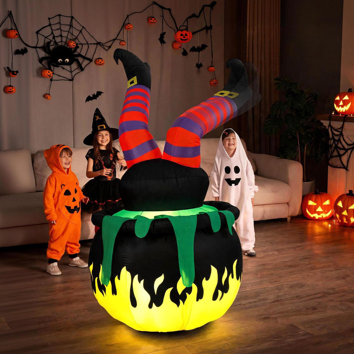 Inflatable witch legs sticking from a glowing black cauldron as children pose in a Halloween-decorated room.