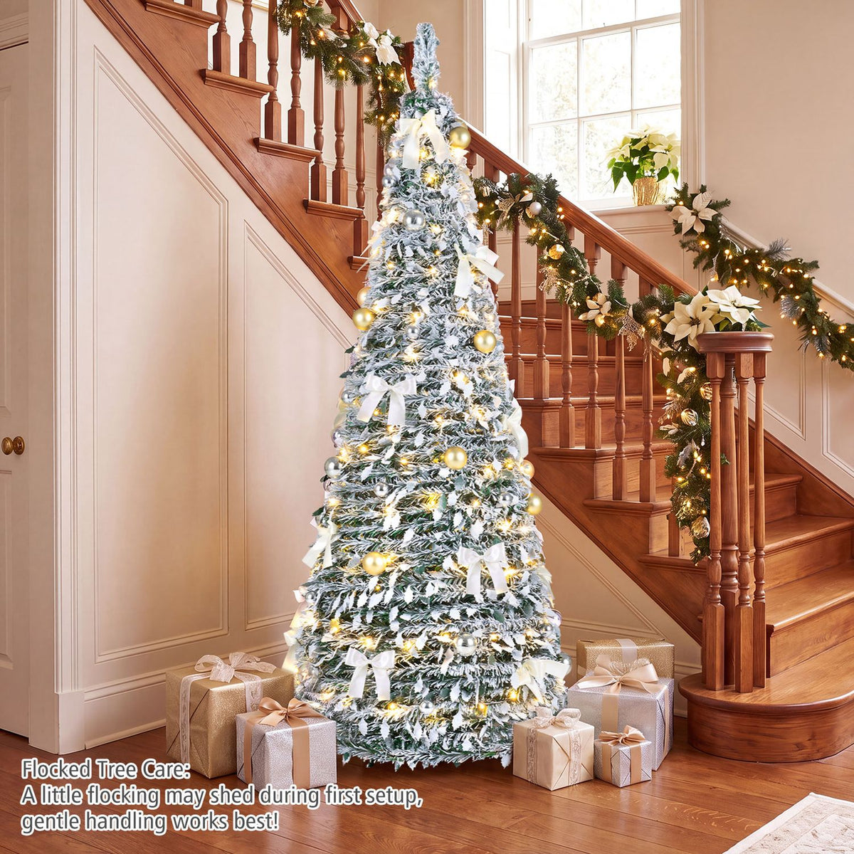 Lit flocked tree decorated with white bows and metallic ornaments in a foyer beneath a staircase.