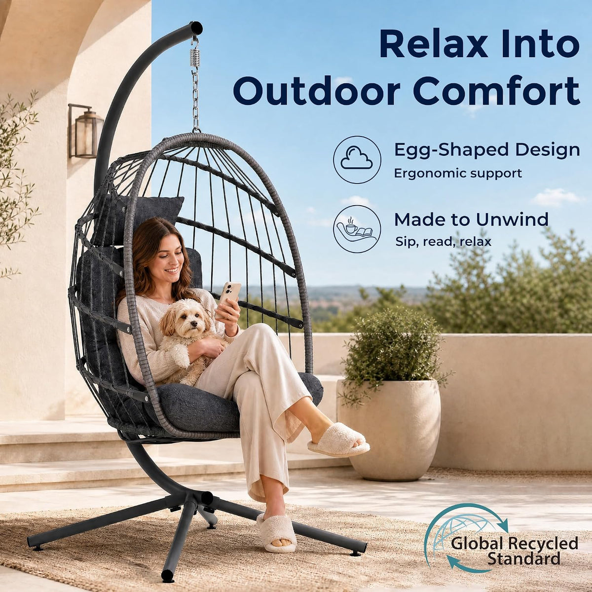 Dark grey wicker hanging chair with cushion and stand on a sunlit patio with a woman holding a small dog