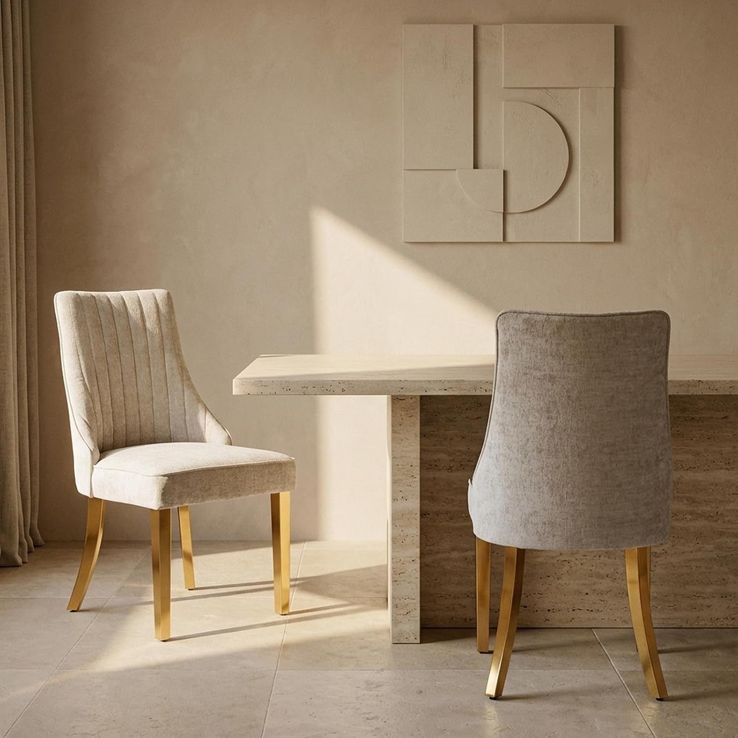 Two beige upholstered dining chairs with gold legs beside a stone dining table in a sunlit room.