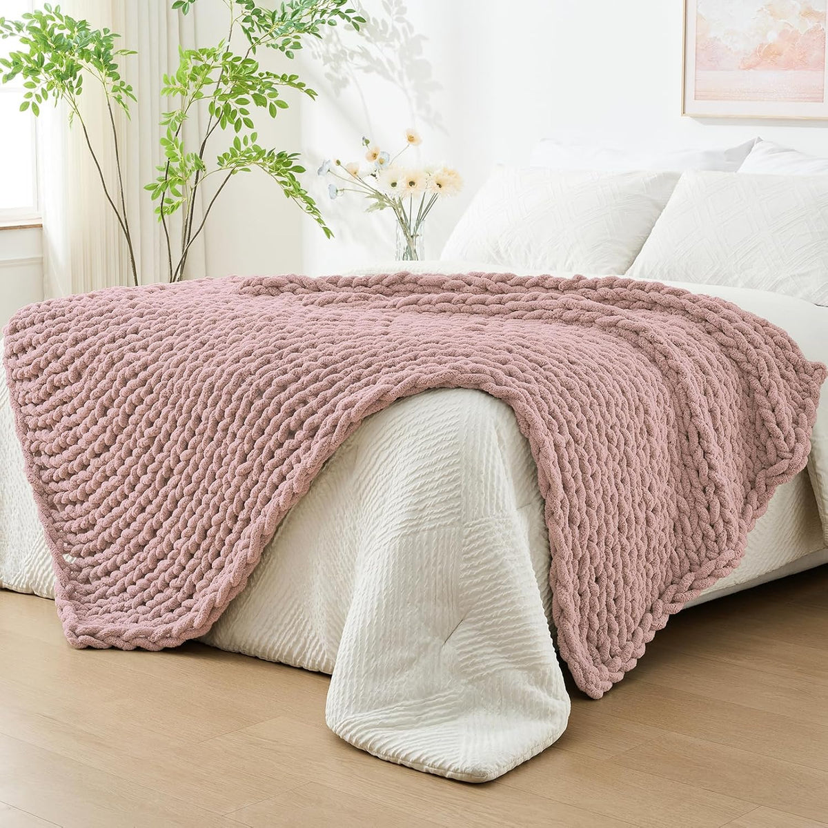 Dusty rose chunky knit throw laid across the foot of a bed with white linens and plants nearby
