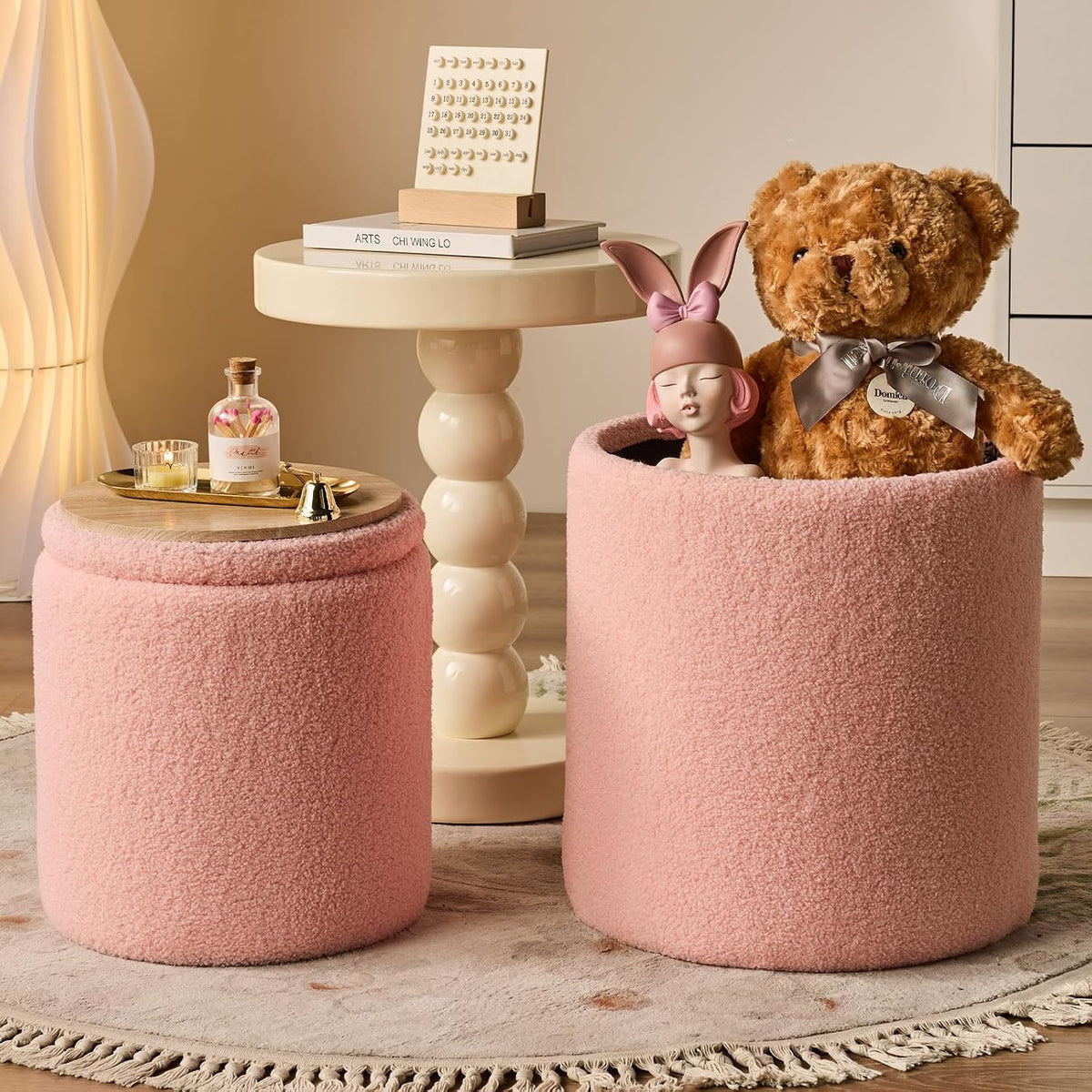 Blush-pink round ottoman pair, one used open as storage holding a teddy and figurine next to a cream pedestal table.