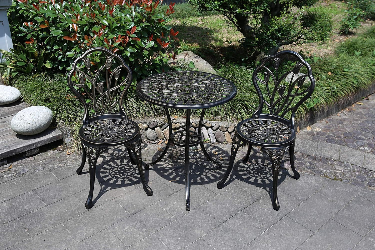 Outdoor bistro set in a garden: round metal table with two matching ornate chairs on stone patio.