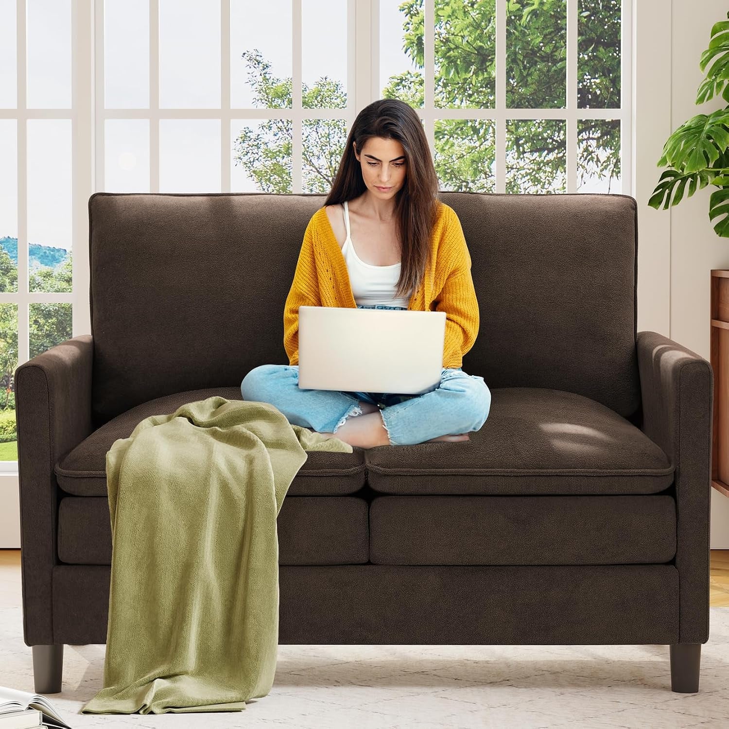 Woman using a laptop while seated cross-legged on a dark brown small loveseat with a green throw.