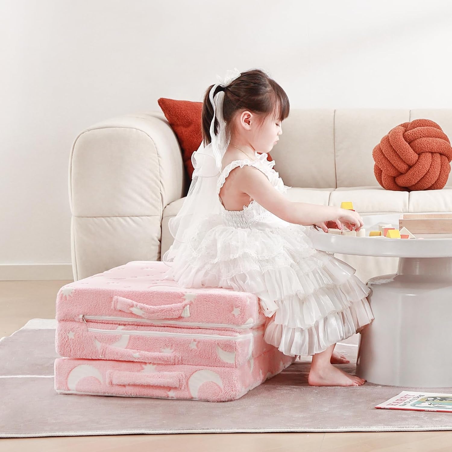Child seated on the folded pink tri-fold mattress beside a low table in a living room.