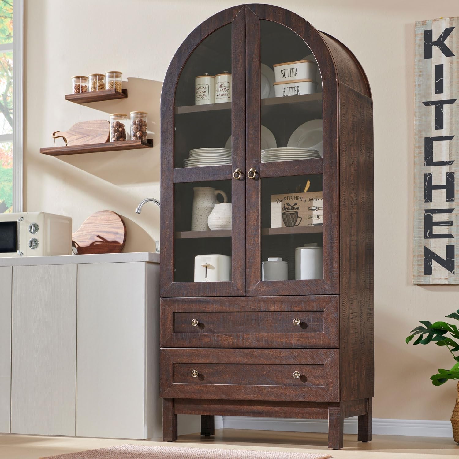 Dark brown arched pantry with glass-front doors and two drawers standing next to a countertop and wall sign.