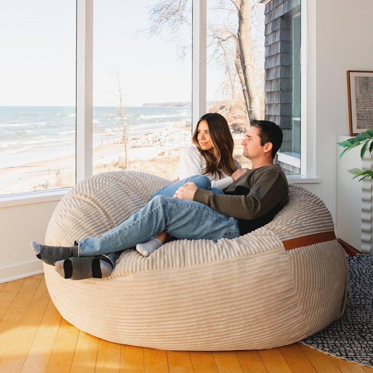 Beige corded-plush oversized lounger with two people seated beside large windows in a lifestyle setting