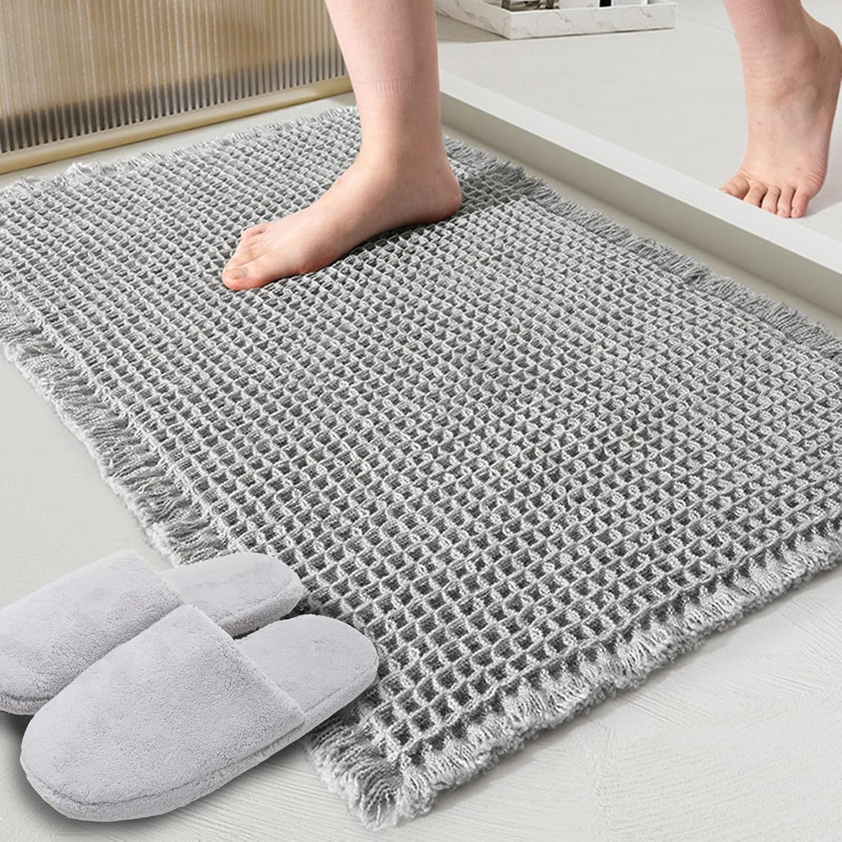 Close-up of a textured light-gray waffle bath mat with a bare foot stepping on it and gray slippers nearby.