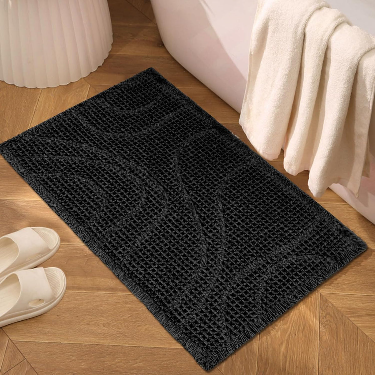 Black textured rectangular mat on wood floor beside a bathtub and towel draped over tub edge.