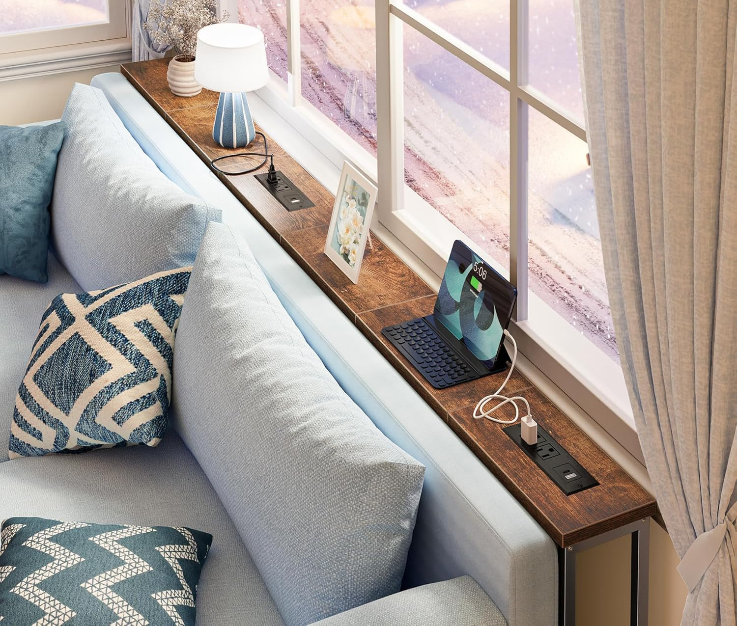 Narrow console table placed behind a sofa by a window, lamp, photo frame and tablet charging on the top