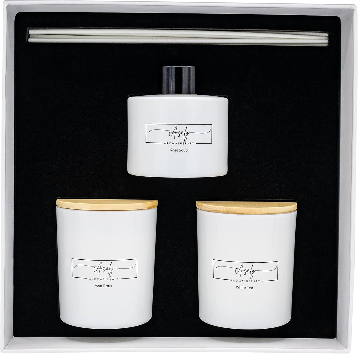 Top-down view of gift box interior showing white reed diffuser bottle, two white glass candles with wooden lids and foam foam