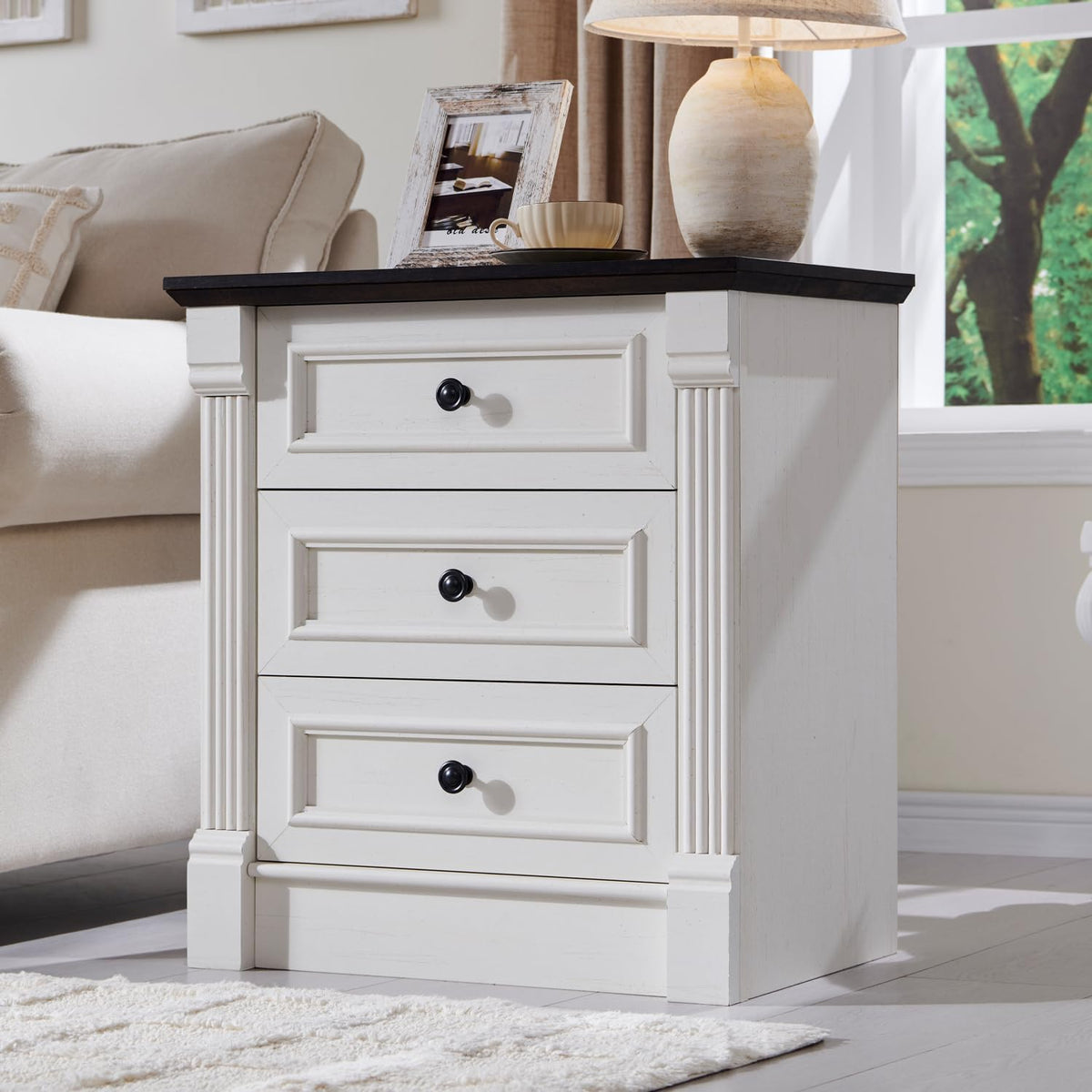 White three-drawer nightstand with dark top and fluted column details beside a sofa, styled with lamp and frame.