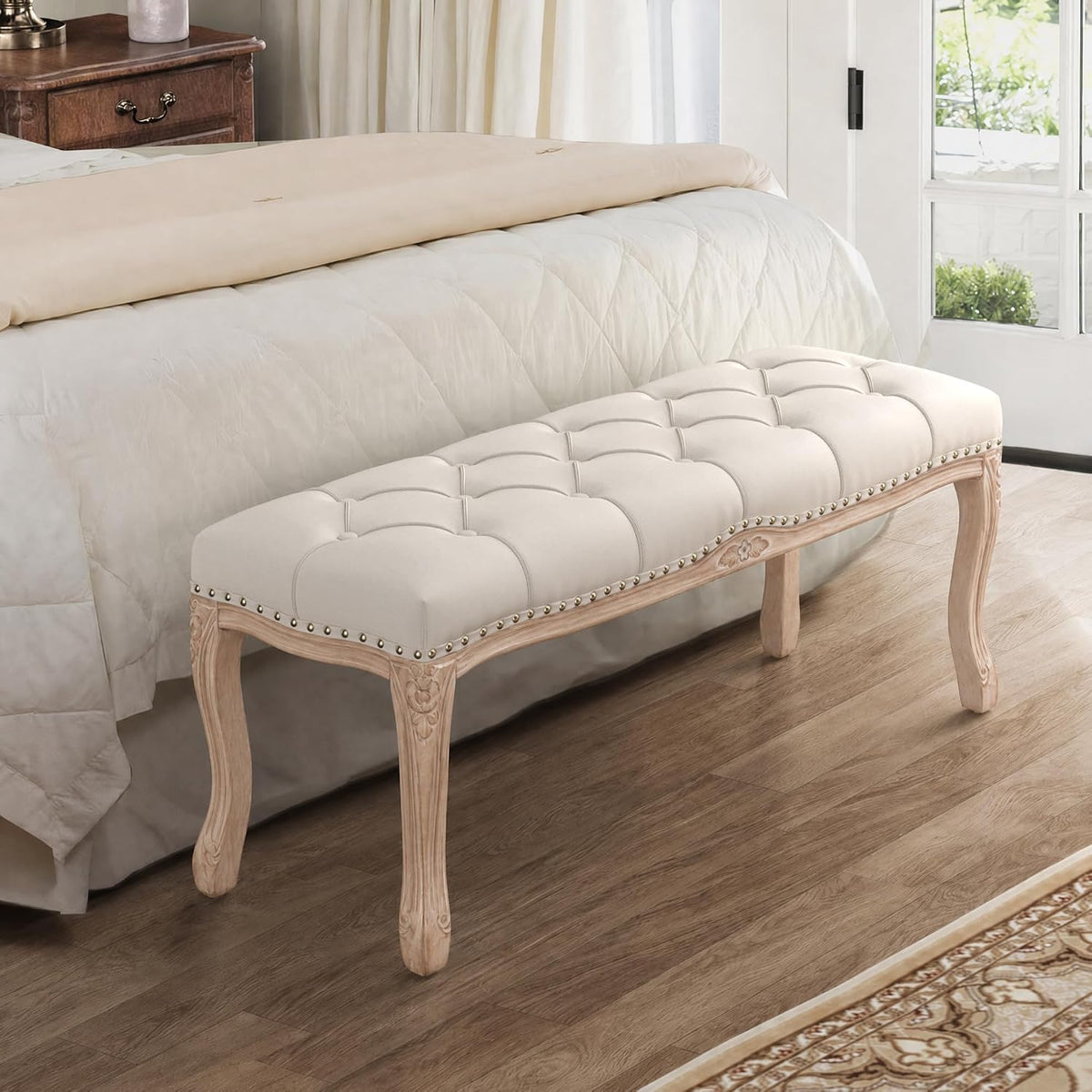 Beige linen tufted bench staged at the foot of a bed in a bedroom setting with carved legs and nailhead trim