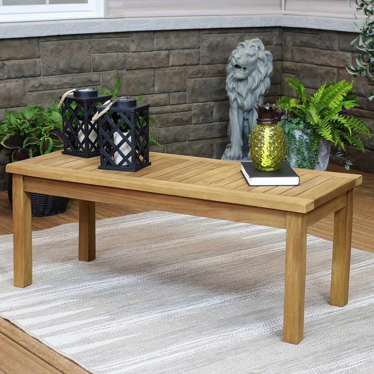 Rectangular light-brown teak coffee table on a striped outdoor rug with lanterns, a book, and a decorative pineapple