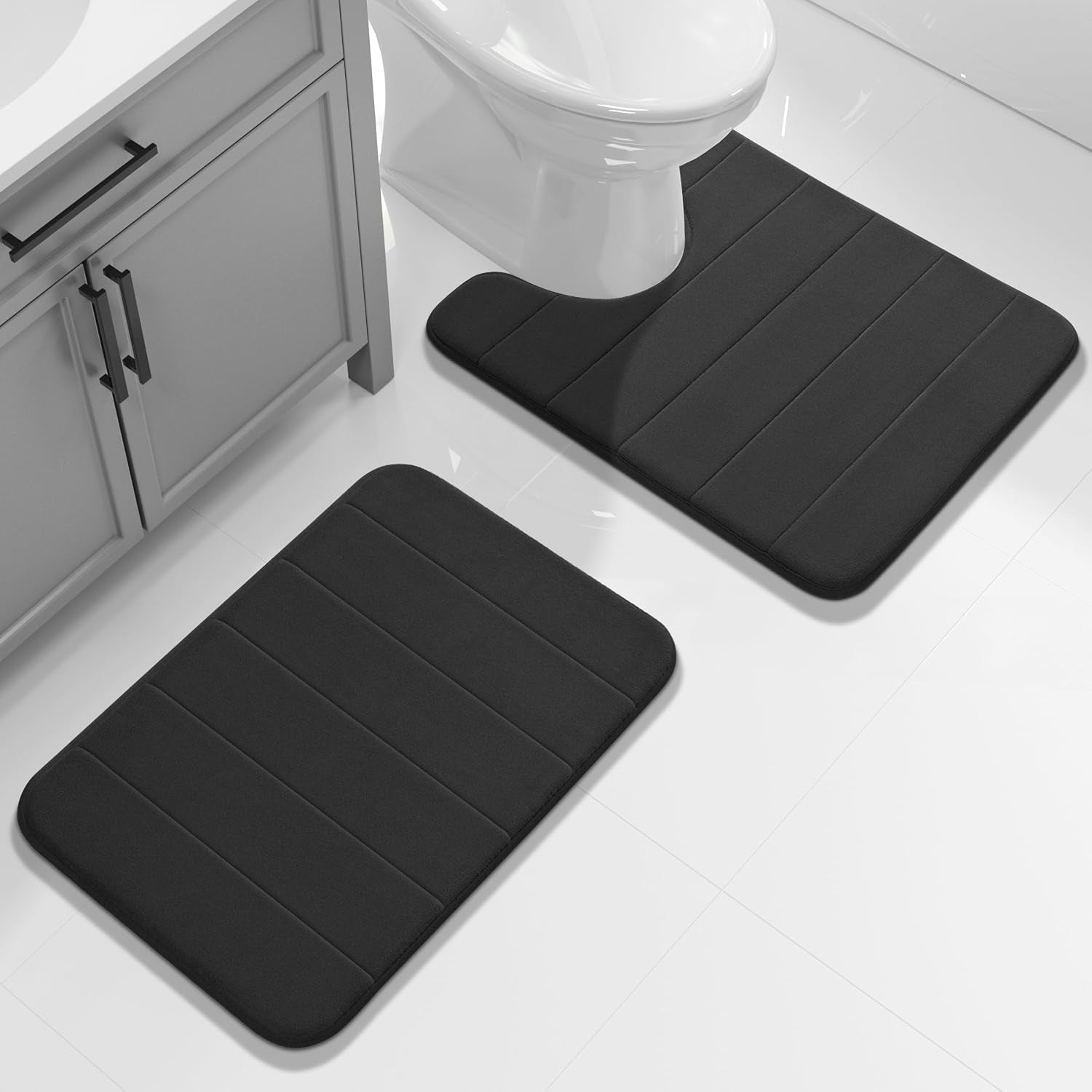 Two black bath mats shown beside a toilet and vanity on a white tiled bathroom floor