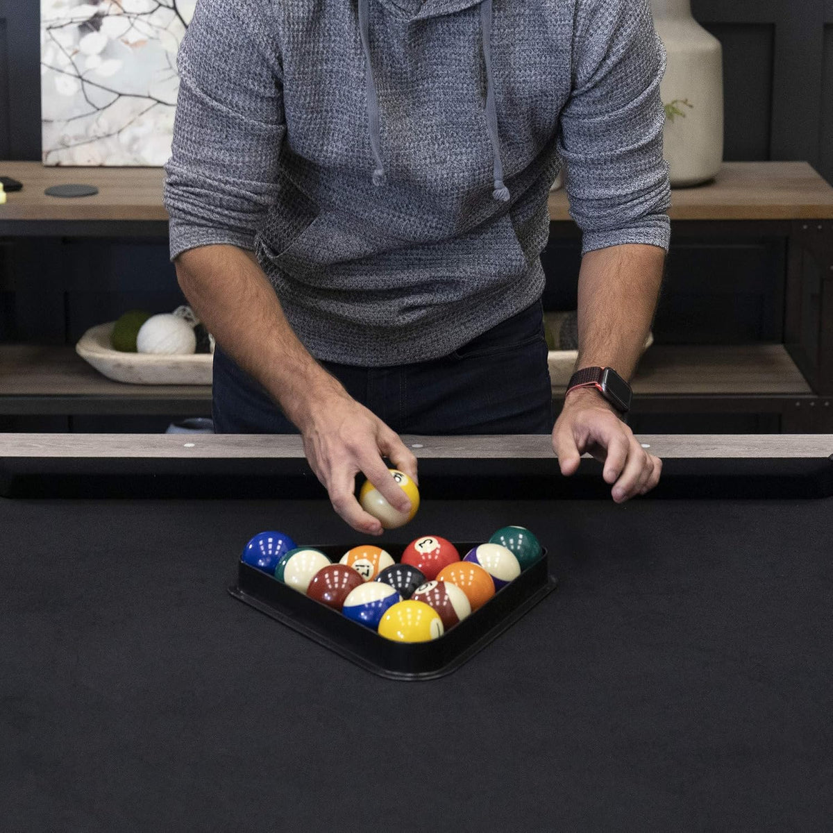 Person racking billiard balls on a black-felt table with a rustic wood rail and shelving in background