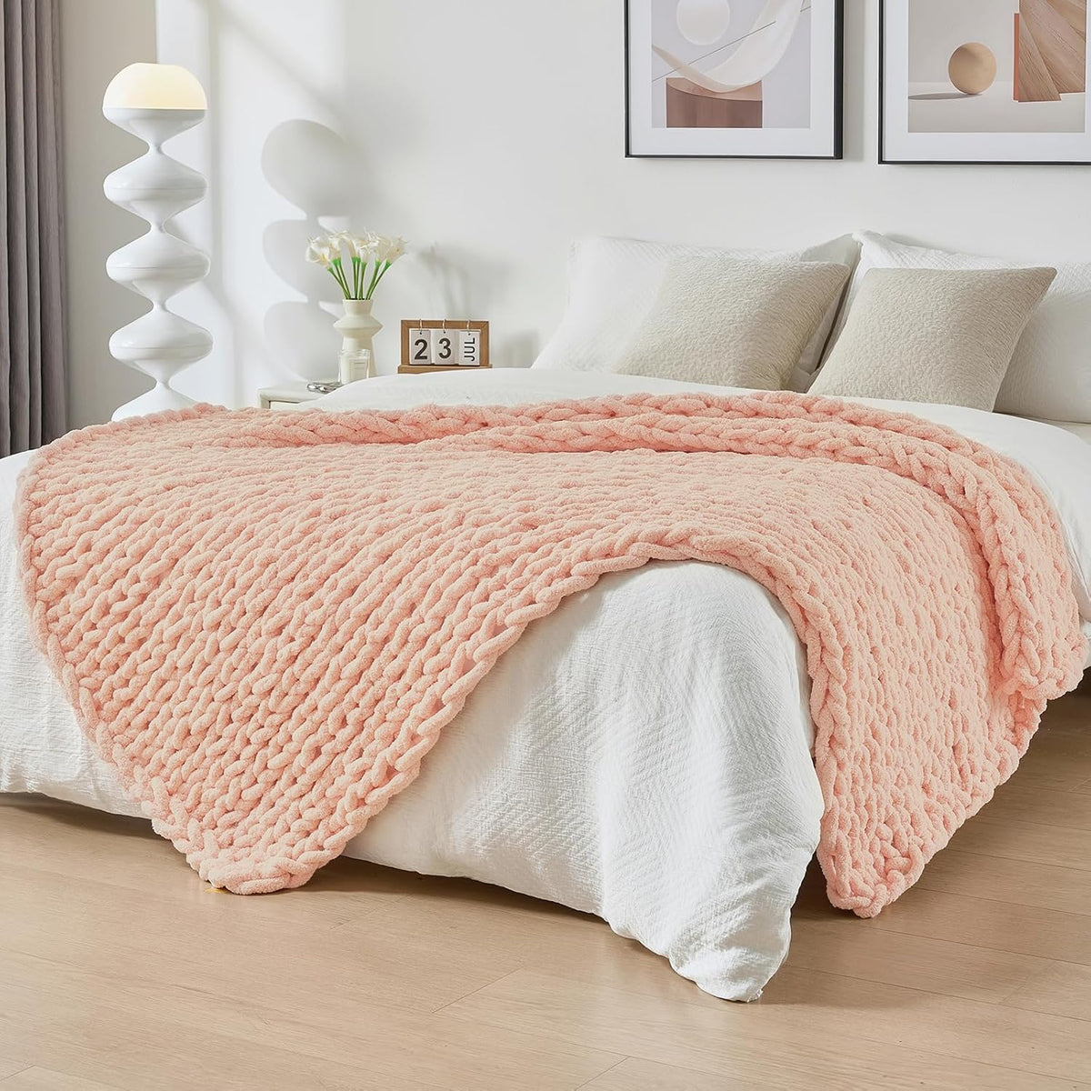 Blush-pink chunky knit throw spread across the foot of a made bed with pillows and modern wall art above.