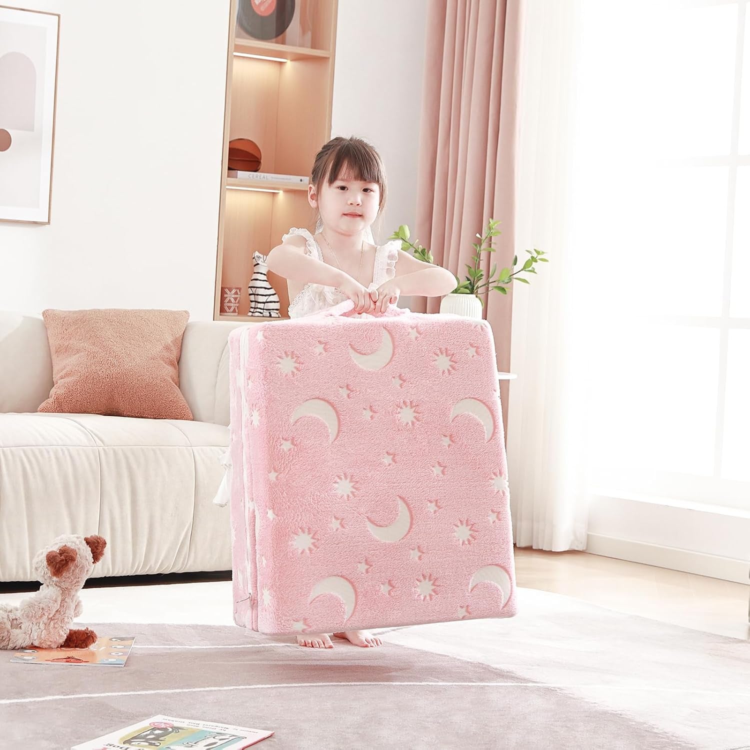 Child standing and lifting the folded pink mattress by its handle in a bright living room.
