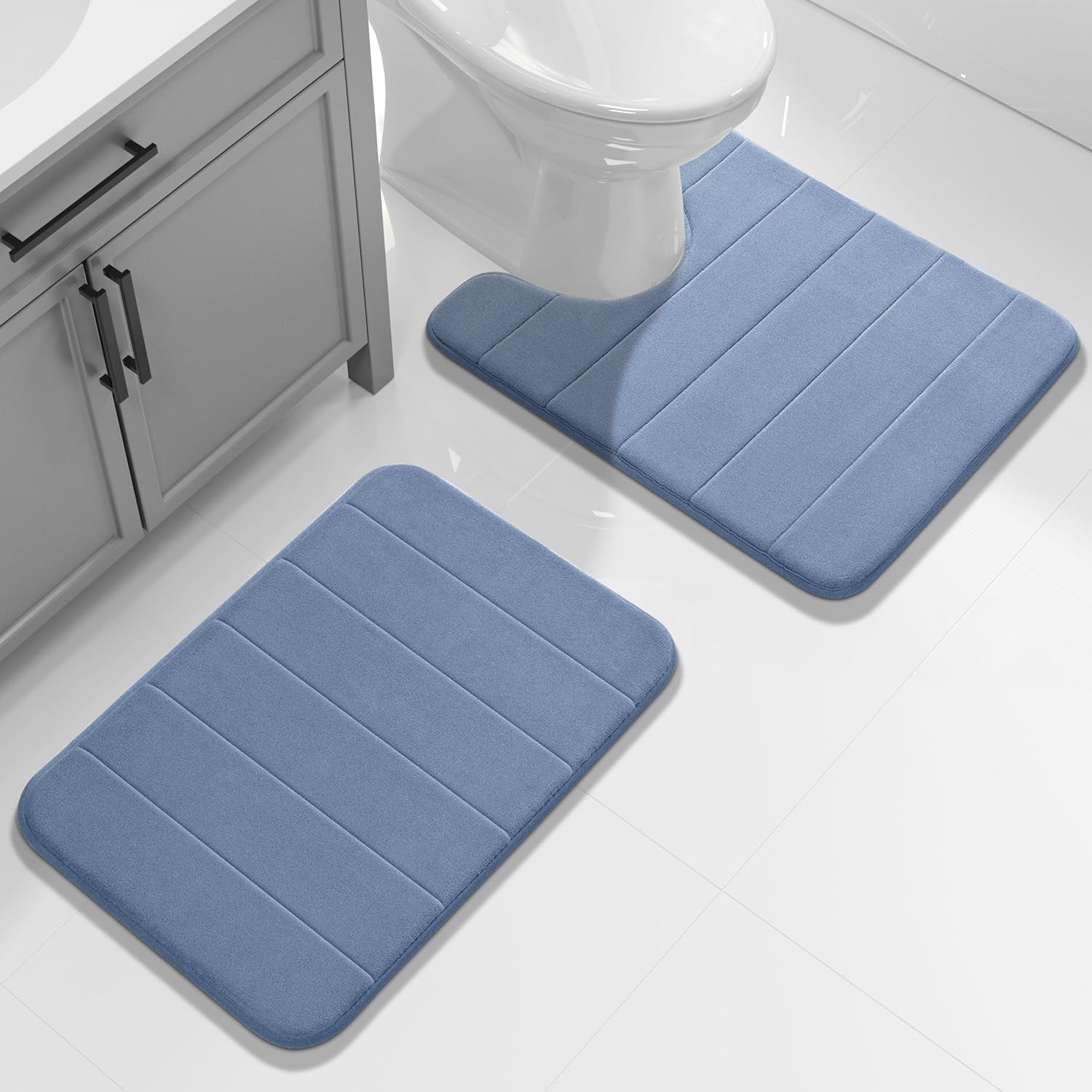 Light blue bath mats placed by a toilet and sink cabinet on white tiled floor.