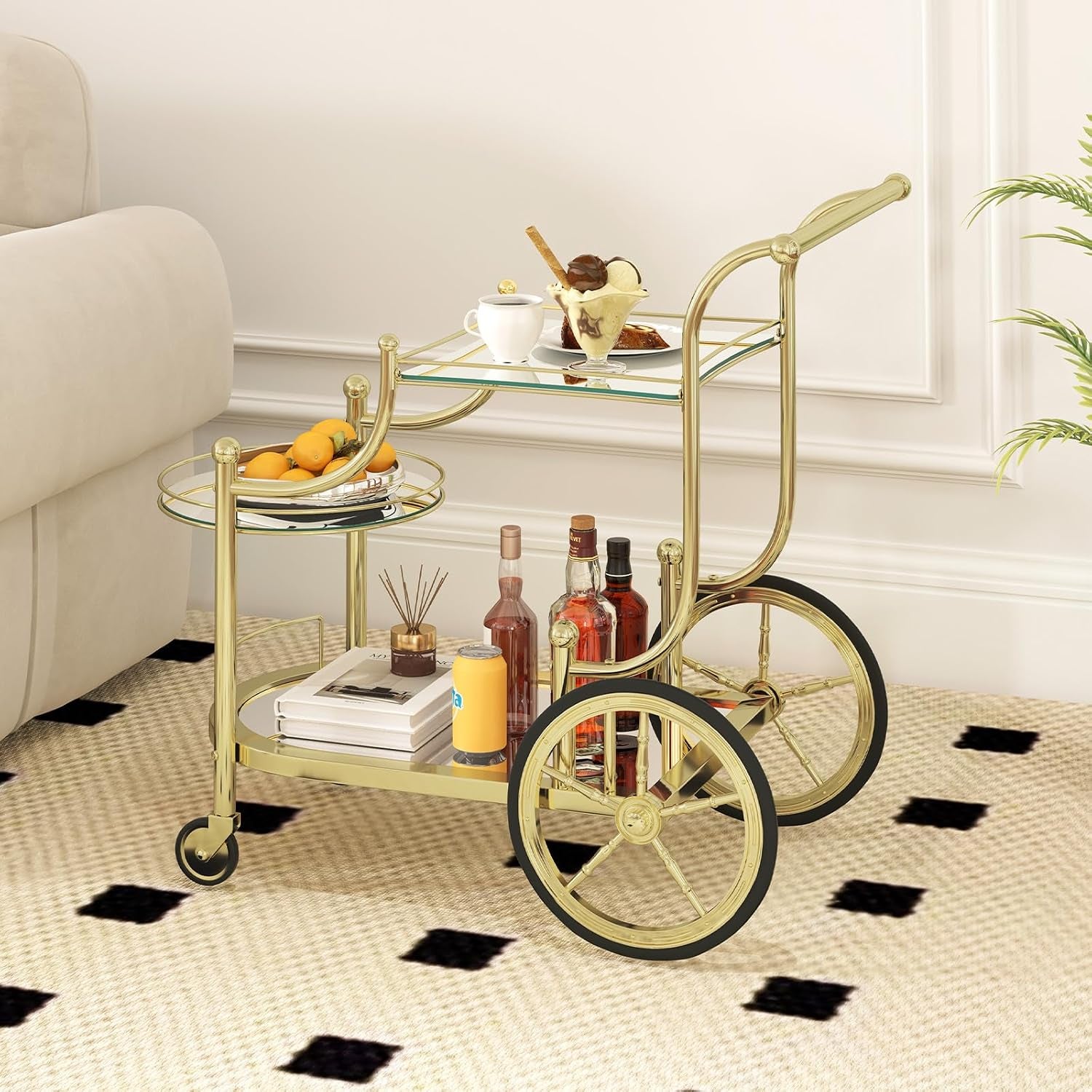 Gold three-tier bar cart on a patterned rug next to a sofa stocked with bottles, books and fruit
