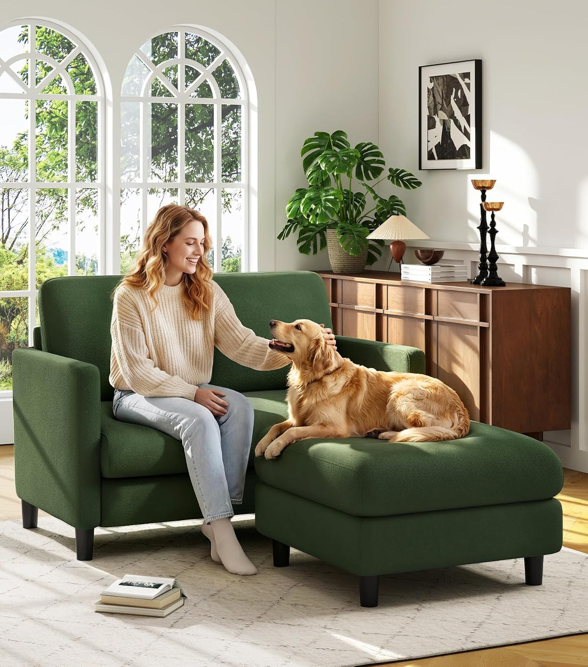Woman smiling while petting a dog on a green loveseat with attached ottoman in a bright living room.