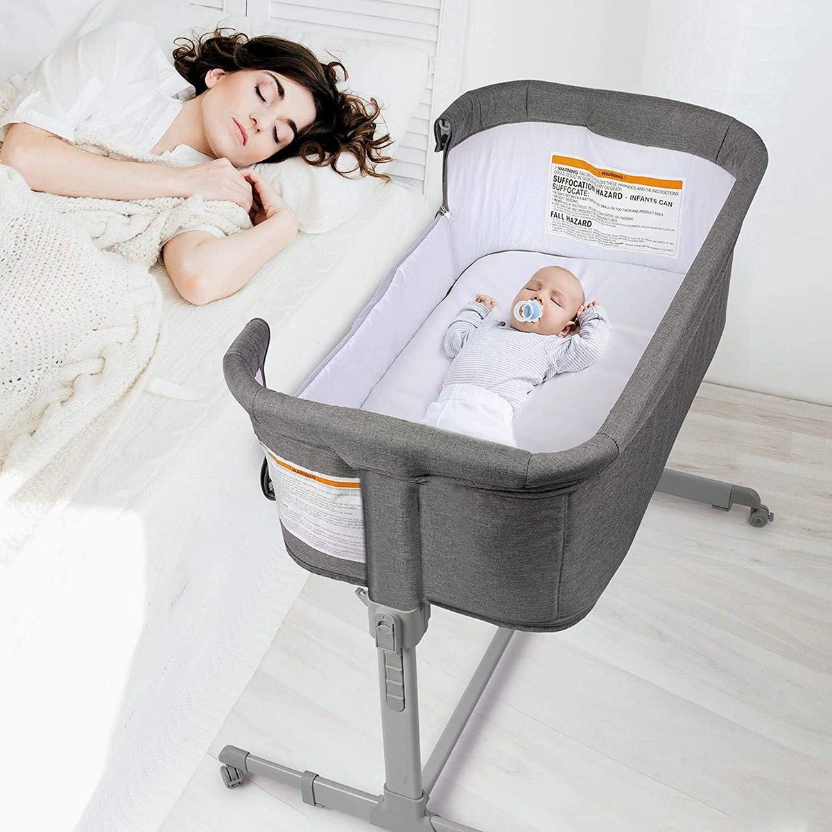 Overhead view of a grey bedside bassinet next to a bed; a sleeping baby lies inside while an adult sleeps in the adjacent