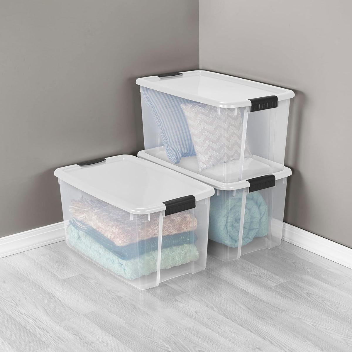 Three clear plastic storage boxes with white lids stacked in a corner holding pillows and blankets.