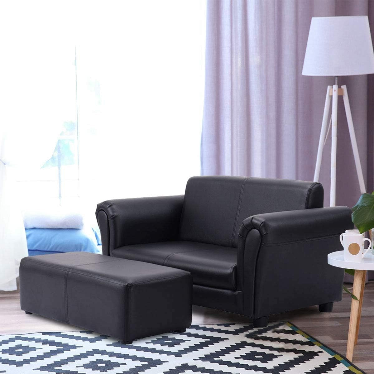 Black two-seat sofa and ottoman staged in a bright living room with patterned rug, floor lamp, and side table.