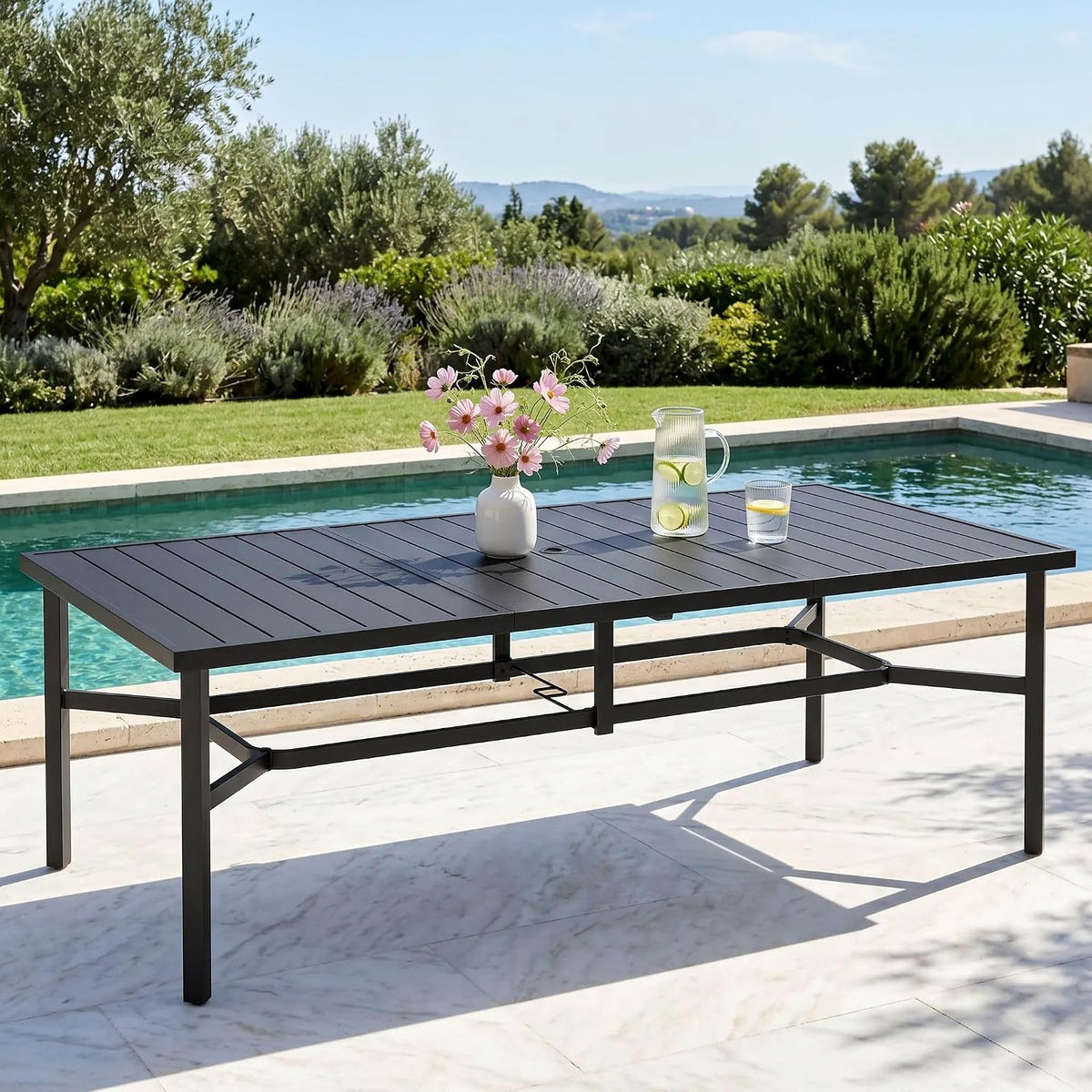 Table placed poolside on a patio with a vase of flowers and a pitcher on top, garden in background.