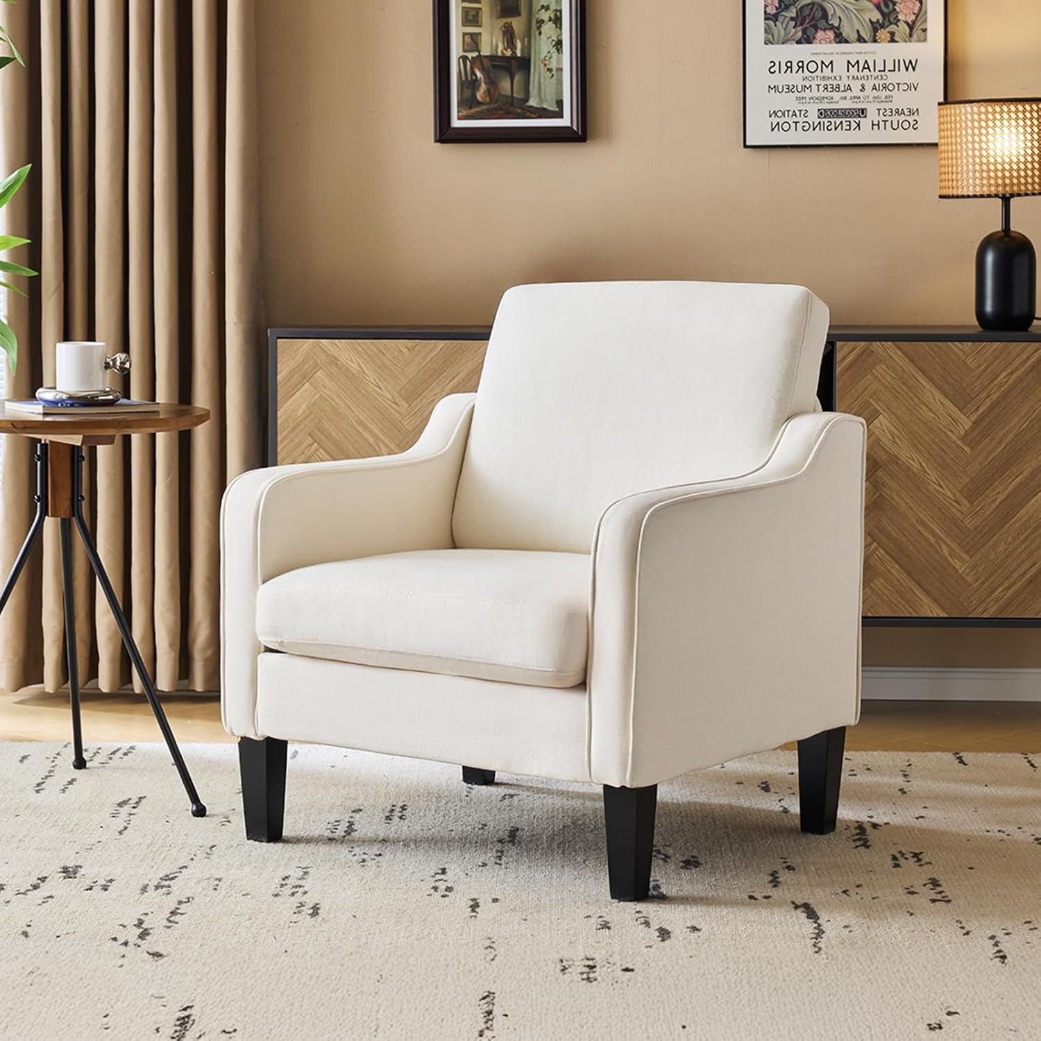 Cream upholstered accent chair in a living room setting with a side table, lamp, rug, and herringbone console behind it.