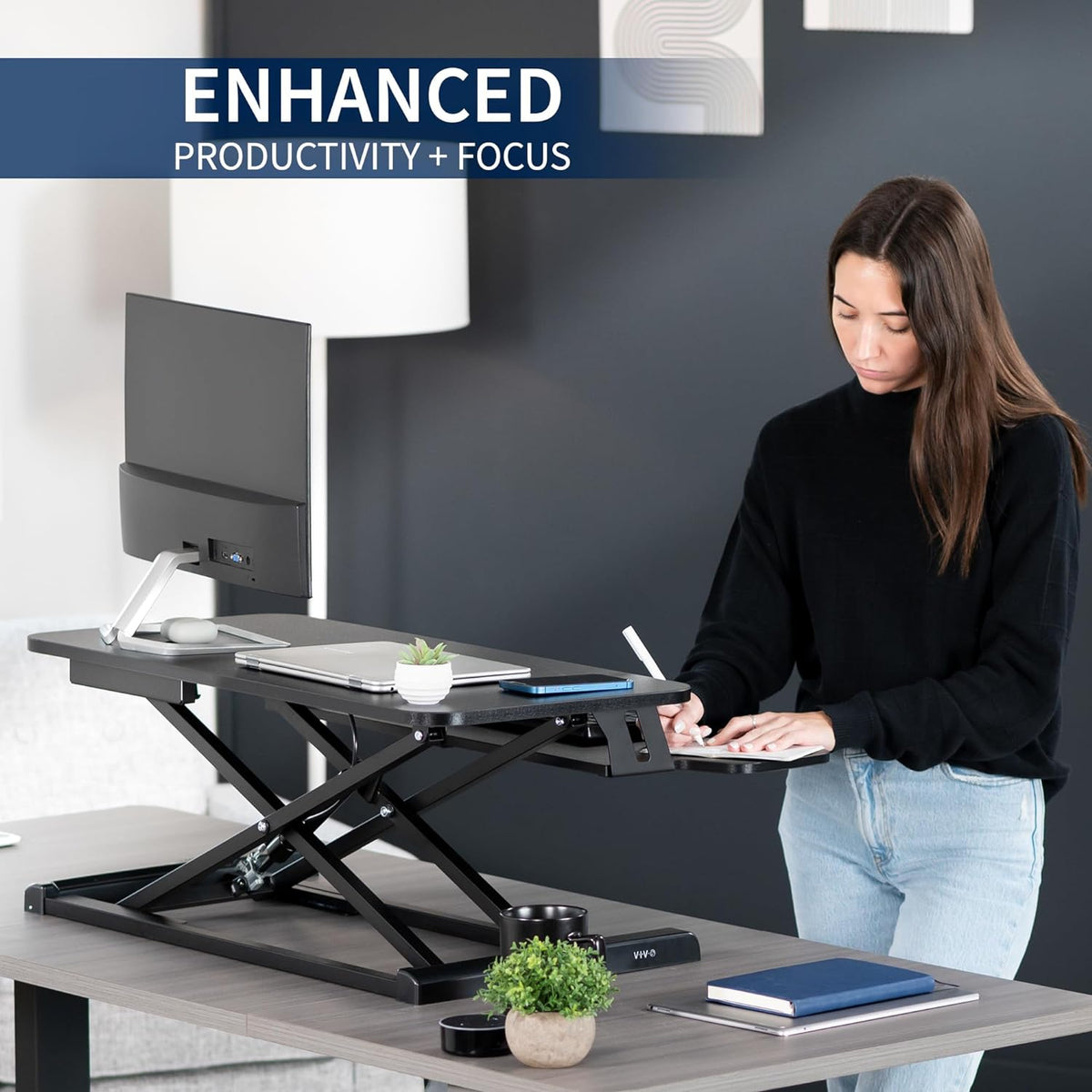 Person standing and writing at a raised black desk converter equipped with a monitor, laptop and desk accessories.