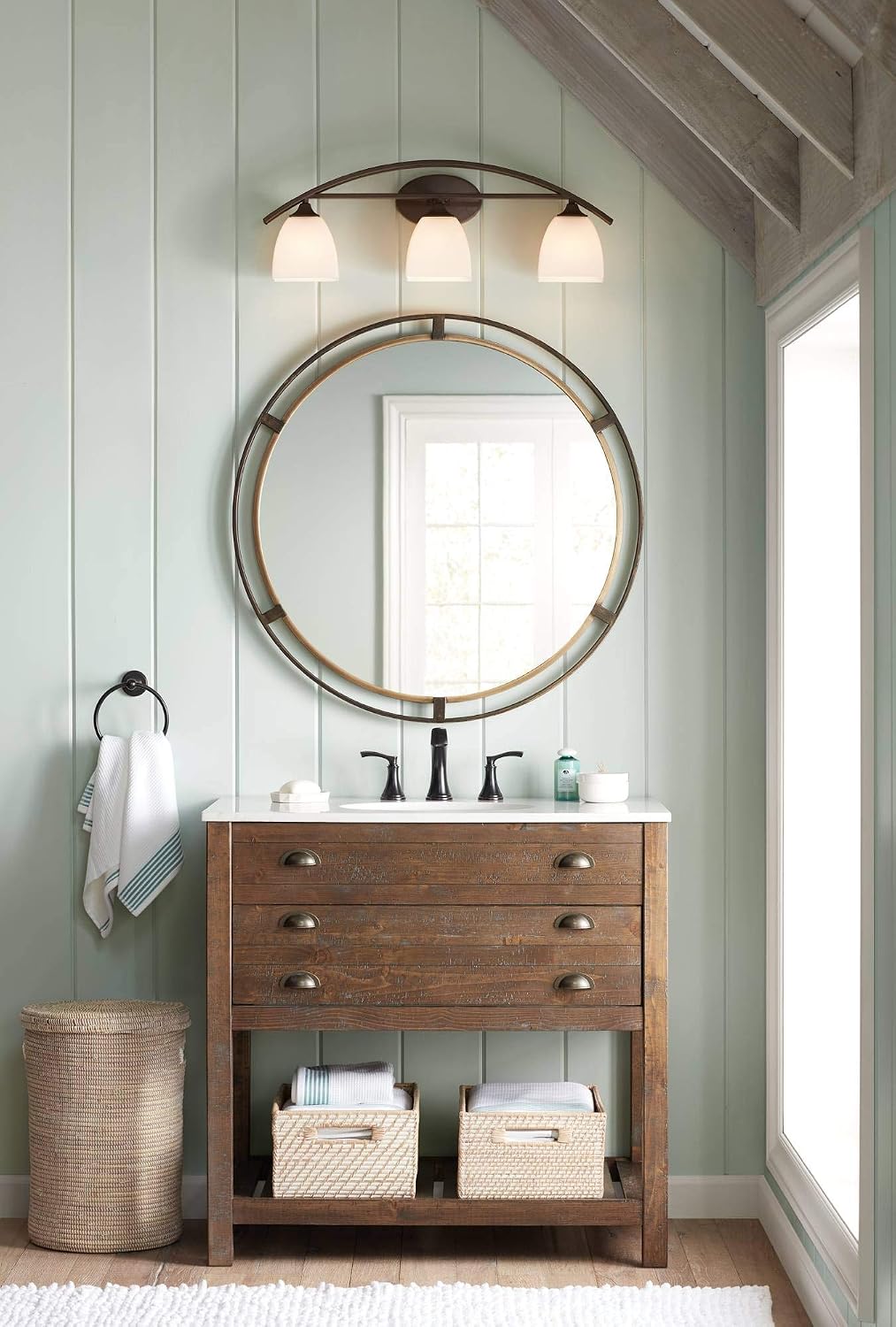Round mirror hung above a rustic wood vanity in a pale green paneled bathroom, lit by a three-light sconce.