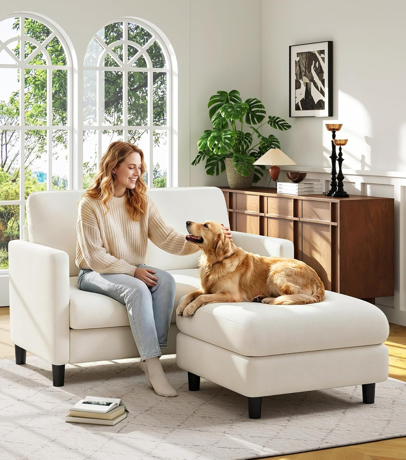 Same scene with the woman and dog on a light cream loveseat and matching ottoman beside a wooden sideboard.