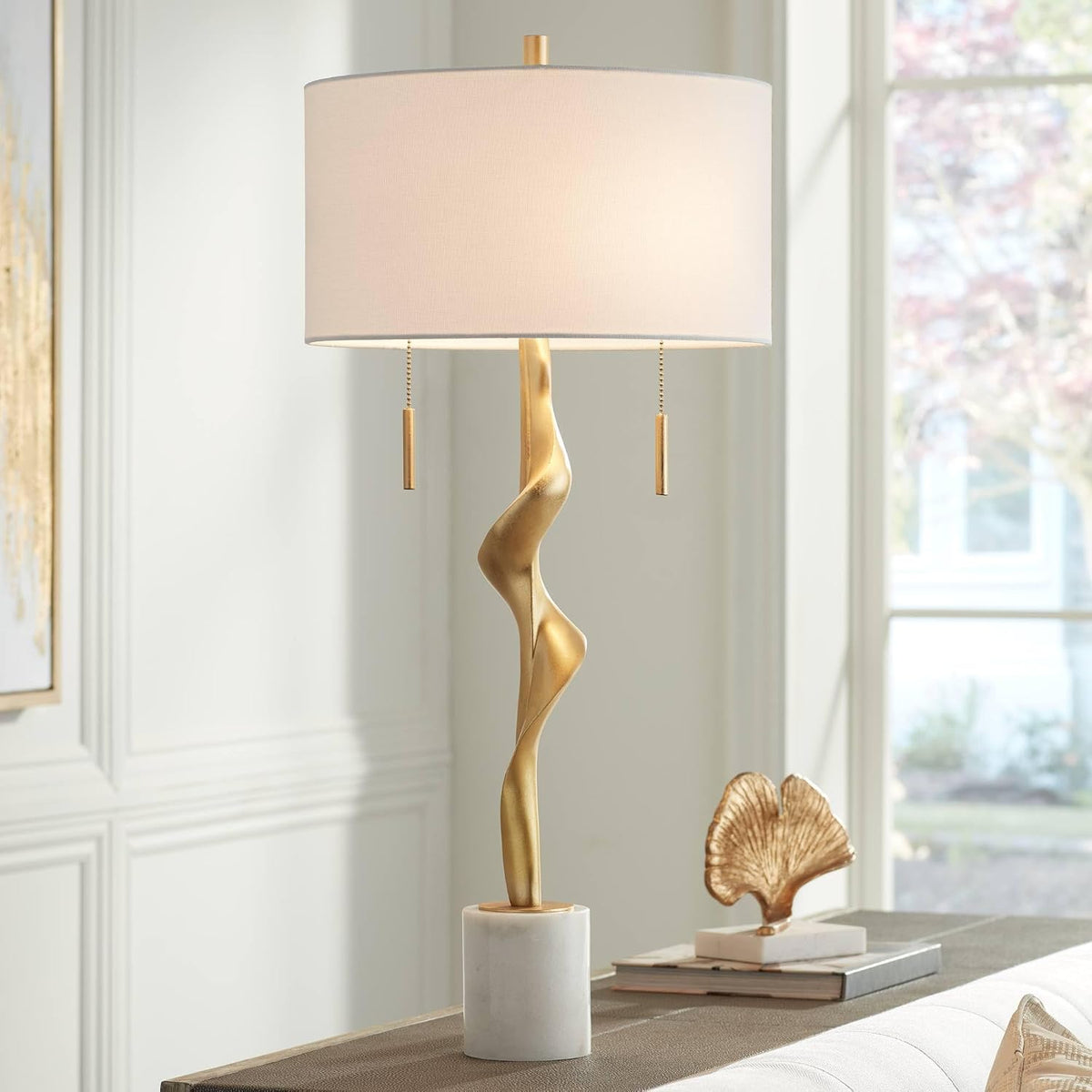 Gold sculptural table lamp with off-white drum shade on a white marble cylinder base, angled front view in a room