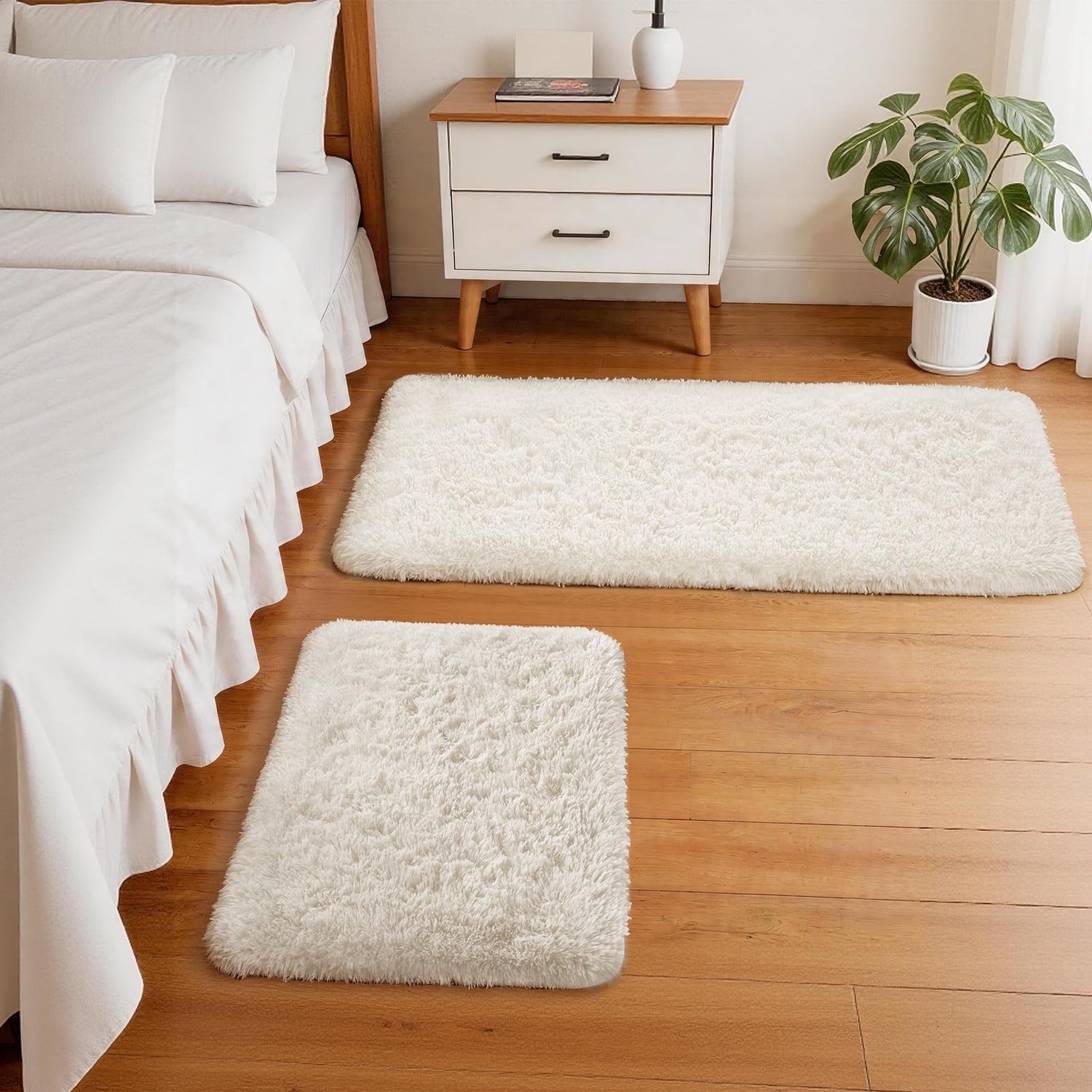 Two cream rectangular shag rugs on a wood bedroom floor next to a bed and a white two-drawer nightstand