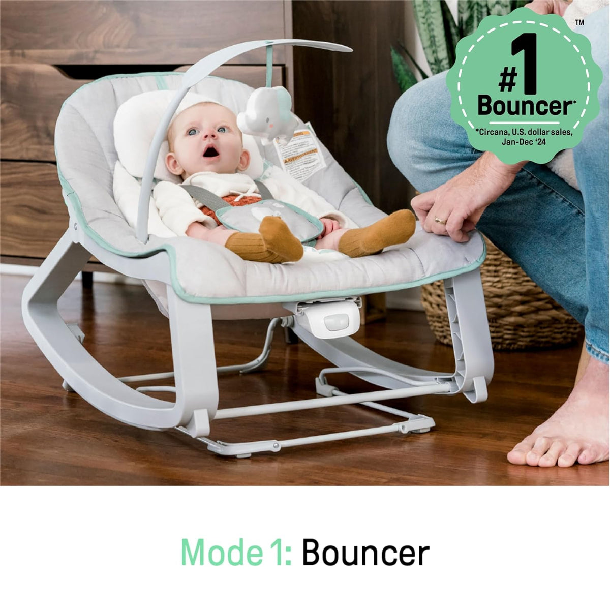 Infant reclined in the padded bouncer on a wooden floor with an adult's hand nearby