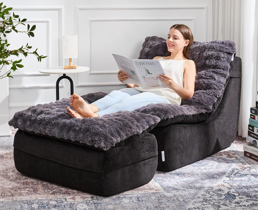 Woman reclining on a dark gray fluffy floor sofa and ottoman, reading in a bright room with side table and rug.