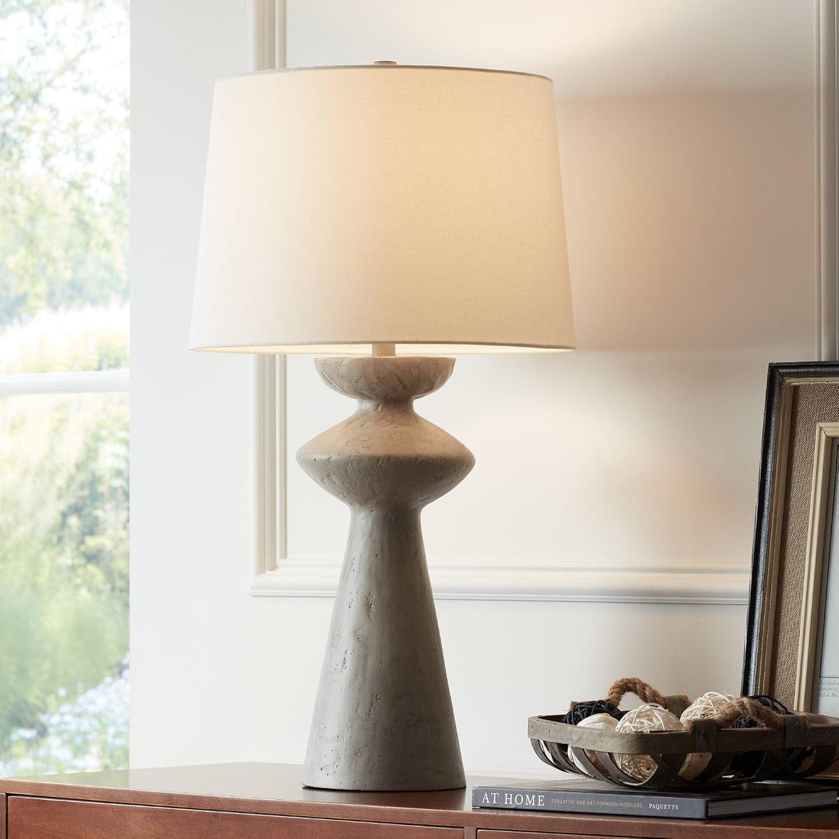 Grey sculptural table lamp with white drum shade on a wooden console in a softly lit room